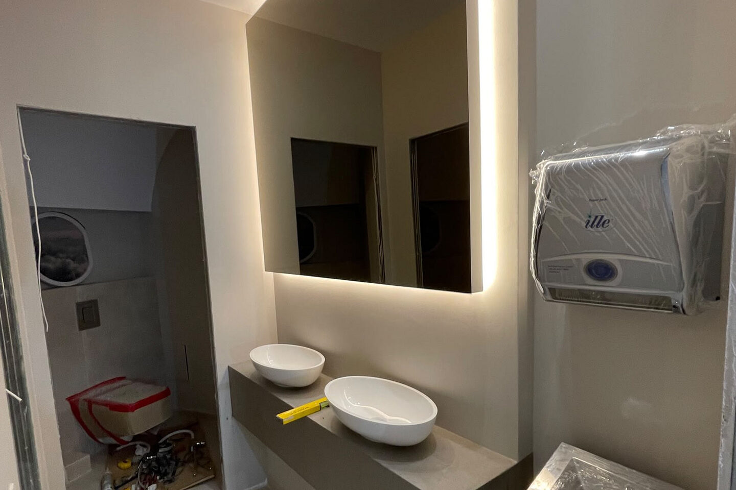 Washroom during fit-out: two basins set on the counter, a backlit mirror and a dispenser still in protective film.