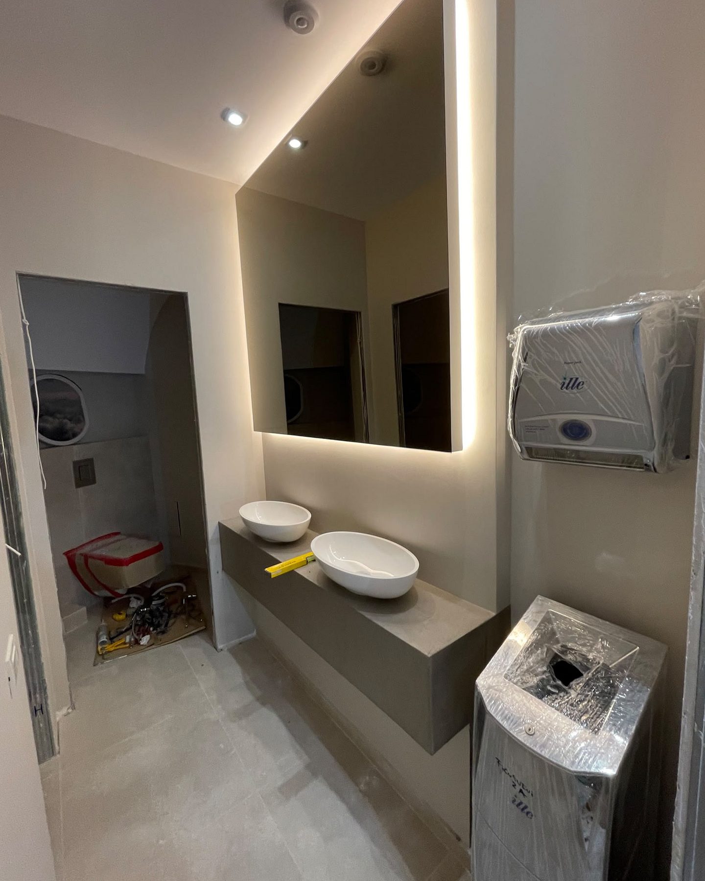 As found: washroom during fit-out, basins set and the mirror lit, fittings still in film.