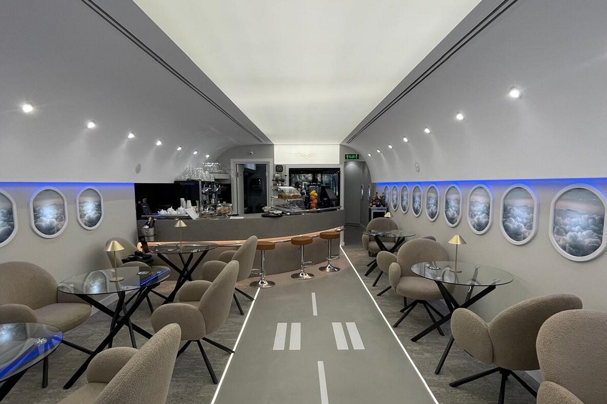 The finished room down its centreline: runway markings on the floor, backlit oval windows along both walls, armchairs and glass tables.