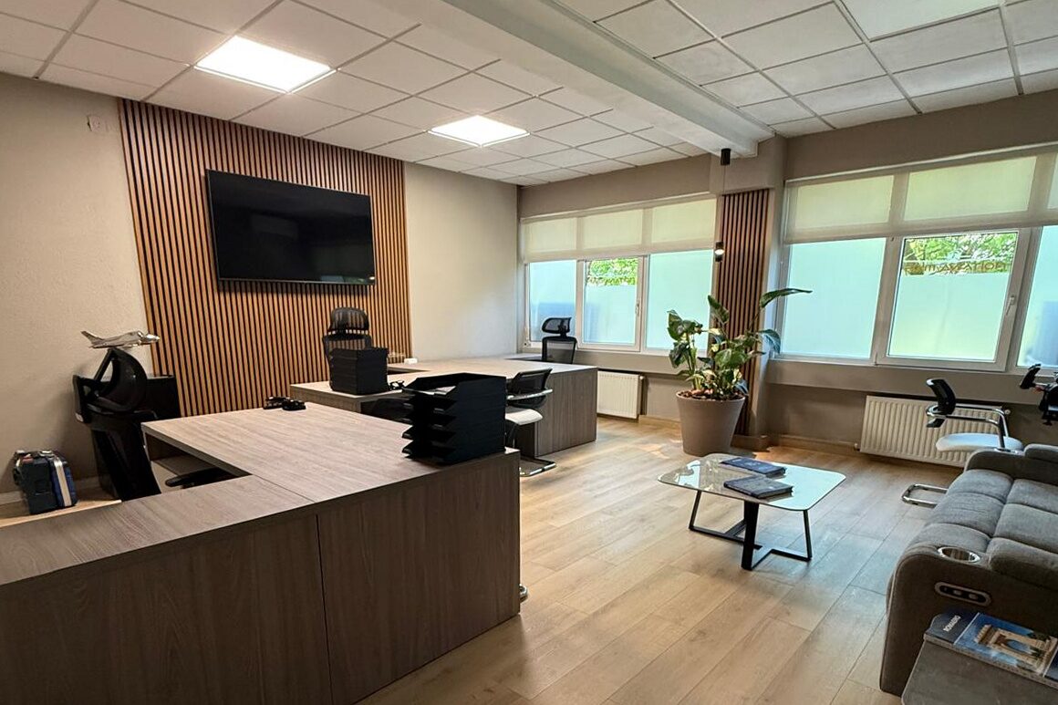 Open-plan office from the entrance: oak-veneer reception counter in the foreground, seating and planting beyond.